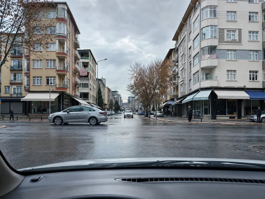 Araç içinden ön cam görünümü: yağmur sonrası ıslak şehir kavşağı, soldan geçen bir otomobil, iki yandaki apartman sıraları karenin kenarlarına kadar giriyor